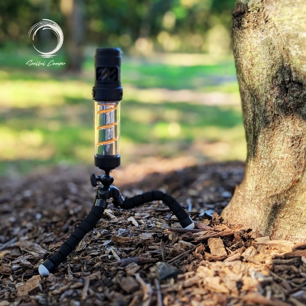 Flexible tripod mount holding camping lantern steady on uneven bark-covered ground beside a tree trunk.