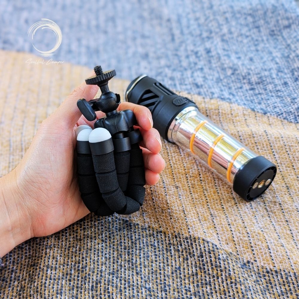Hand holding FlexiMount tripod beside Silent Lantern on a textured camping mat