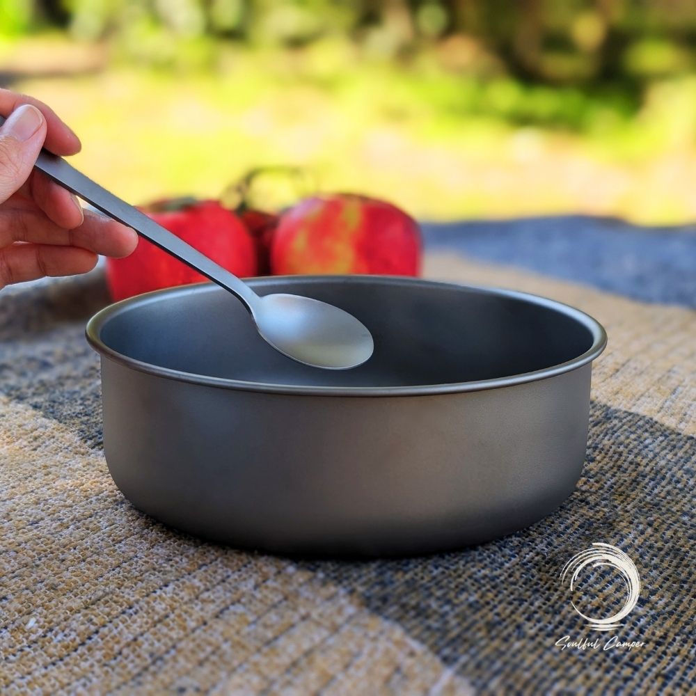 Hand holding spoon over titanium camping bowl with apples in background