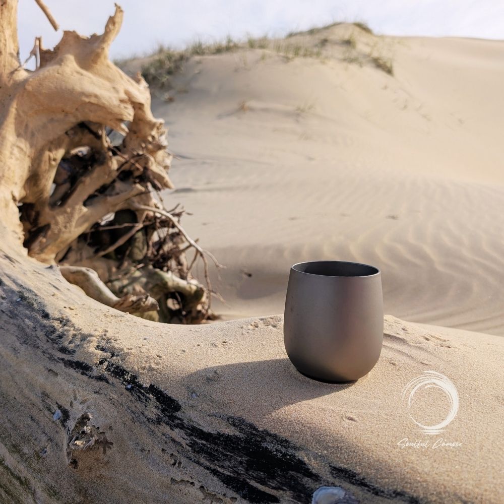 Titanium cup sitting on driftwood in natural sand dune setting, perfect for eco-conscious campers.