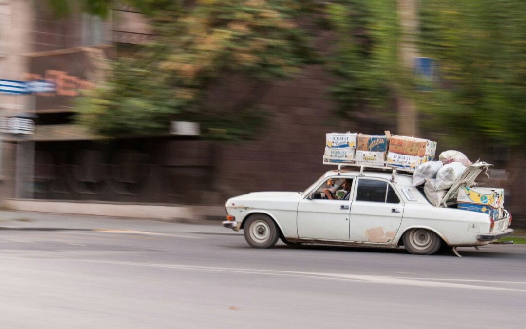 A white vintage car overloaded with boxes and bags strapped to the roof, driving fast down a city street, symbolizing the weight of carrying too much.