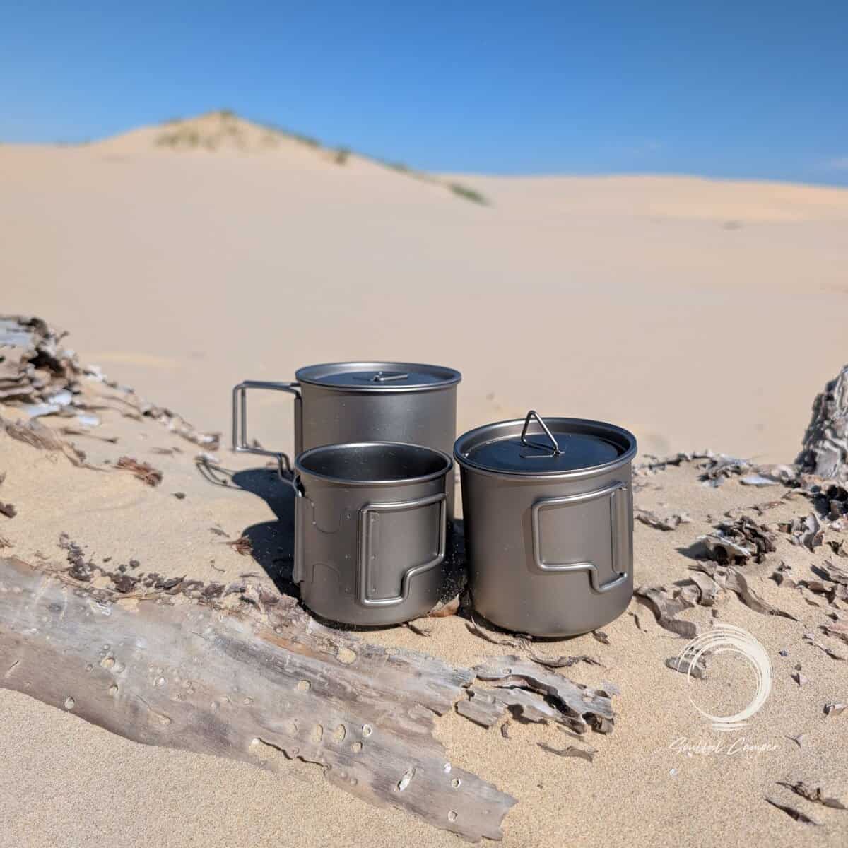 Titanium camping mugs in three sizes from the Ti-3 Mug Stack displayed on driftwood in the Anna Bay sand dunes, showing lightweight nesting outdoor mug set.